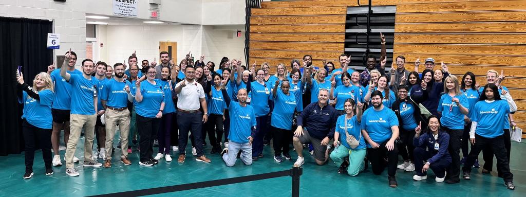 A large group of people in matching blue shirts pose together inside a gymnasium, smiling and raising their index fingers as a gesture of unity and celebration Some people are kneeling in the front row