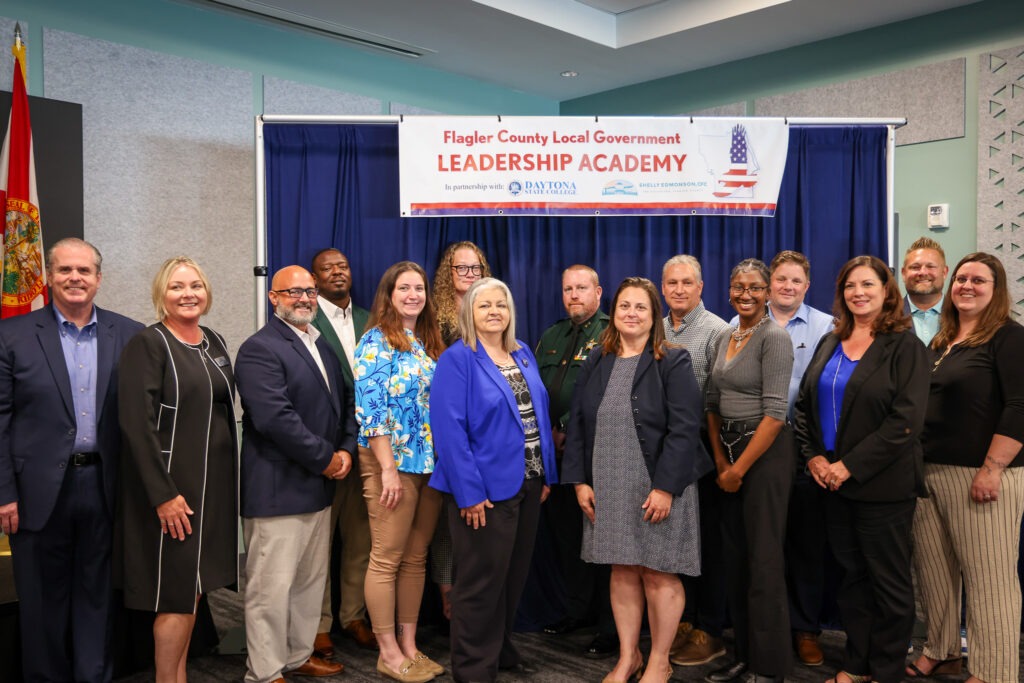 A group of fourteen adults, both men and women, stand smiling in front of a banner that reads "Flagler County Local Government Leadership Academy" inside a conference room A group of fourteen adults, both men and women, stand smiling in front of a banner that reads "Flagler County Local Government Leadership Academy" inside a conference room