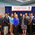 A group of fourteen adults, both men and women, stand smiling in front of a banner that reads "Flagler County Local Government Leadership Academy" inside a conference room