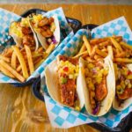 Two baskets lined with blue and white checkered paper, each holding seasoned fish tacos topped with pineapple salsa and a side of French fries, on a wooden table