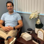 A man sits smiling in a chair next to a table with a potted orchid, stacked books, a candle, and a “DAD” sign Blinds cover the window behind him and the room has a calm, welcoming atmosphere