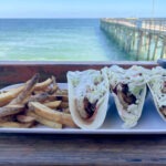 A plate with three tacos filled with grilled fish and slaw, served with French fries, sits on a wooden table overlooking the ocean and a pier on a sunny day