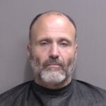 A middle aged man with a beard and receding hairline, wearing a black garment, poses against a plain gray background for a mugshot