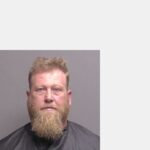 A middle aged man with light skin, short blond hair, and a full blond beard faces the camera against a plain gray background, wearing a dark cape commonly seen in mugshots