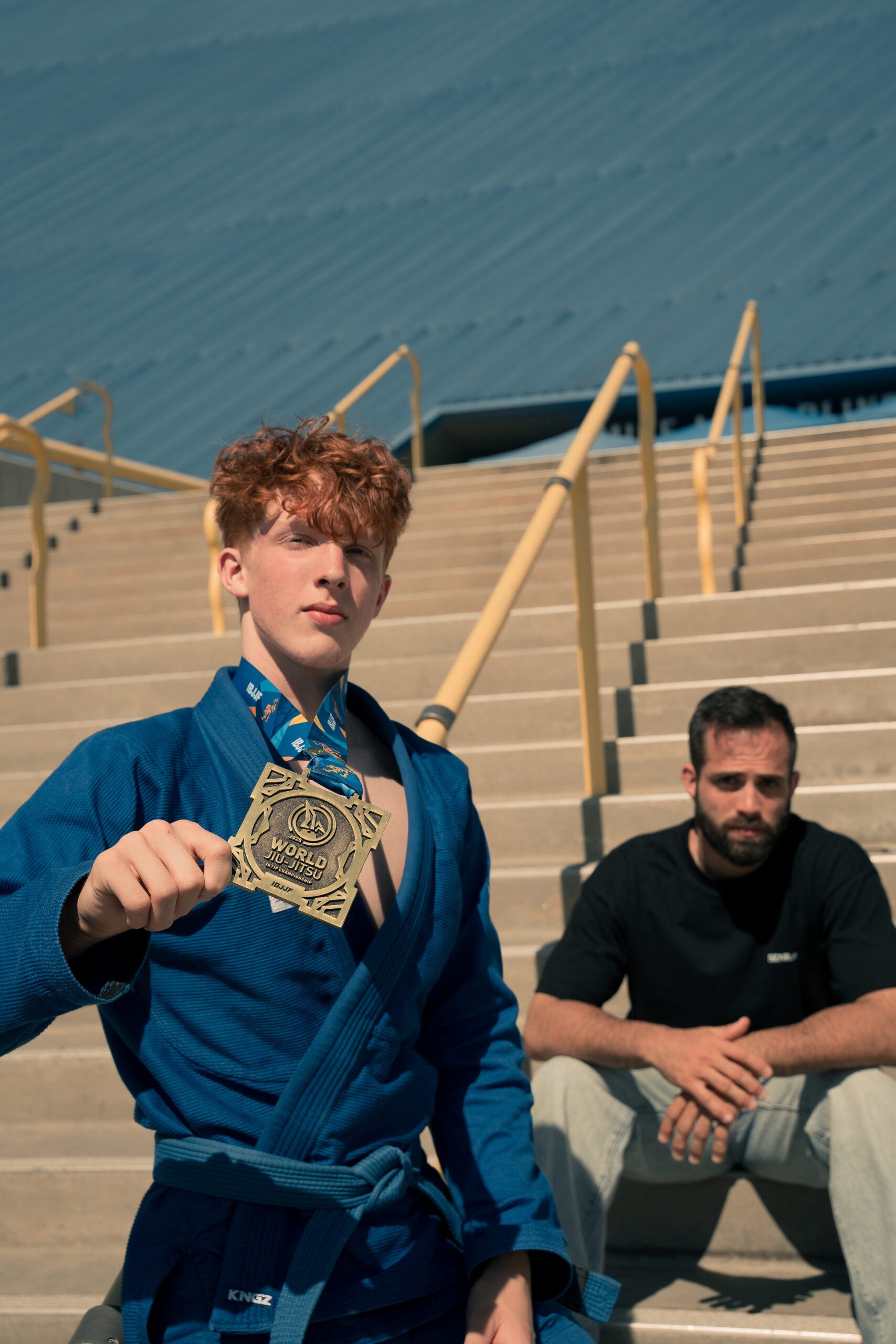 A young man in a blue martial arts uniform proudly holds a gold medal in front of stadium steps, while another man in casual clothes sits on the steps behind him A young man in a blue martial arts uniform proudly holds a gold medal in front of stadium steps, while another man in casual clothes sits on the steps behind him
