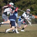 Three youth lacrosse players, wearing helmets and protective gear, are in action on a grassy field One player is being checked by an opponent while another player runs towards them, all focused on the game Trees are in the background