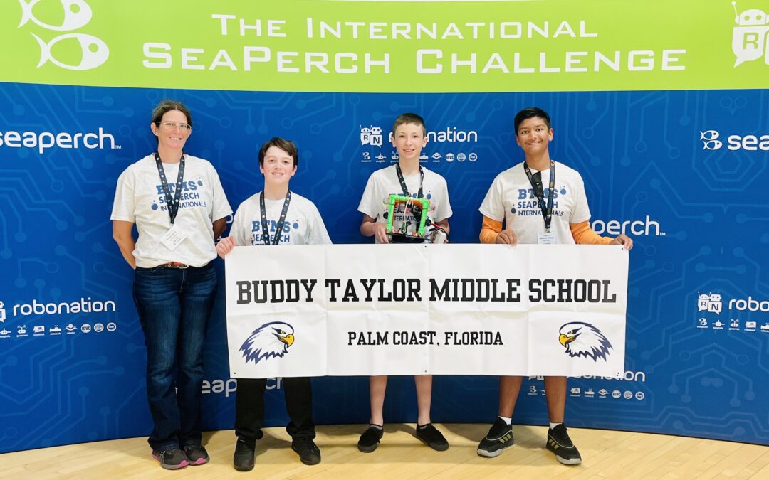 Four people stand in front of a blue backdrop at the International SeaPerch Challenge, holding medals and a robot, with a "Buddy Taylor Middle School, Palm Coast, Florida" banner featuring an eagle logo