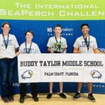 Four people stand in front of a blue backdrop at the International SeaPerch Challenge, holding medals and a robot, with a "Buddy Taylor Middle School, Palm Coast, Florida" banner featuring an eagle logo