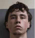 A young man with pale skin, short dark hair, and blue eyes stands against a gray and beige wall, looking directly at the camera with a neutral expression