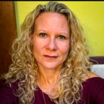 A woman with long, curly blonde hair and light skin looks directly at the camera She is wearing a burgundy top and is in front of a yellow wall