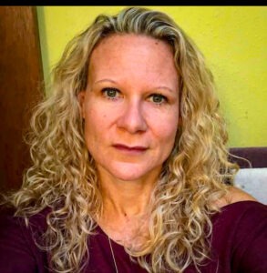 A woman with long, curly blonde hair and light skin looks directly at the camera She is wearing a burgundy top and is in front of a yellow wall