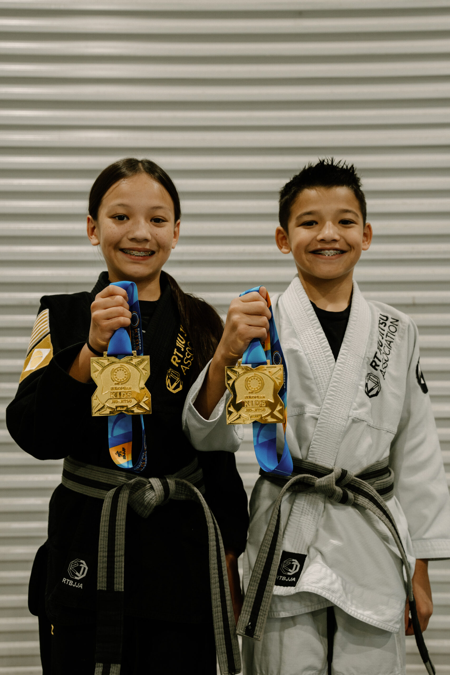 Two smiling children in martial arts uniforms proudly hold up gold medals One wears a black gi, the other a white gi, both with gray belts, standing in front of a corrugated metal background Two smiling children in martial arts uniforms proudly hold up gold medals One wears a black gi, the other a white gi, both with gray belts, standing in front of a corrugated metal background