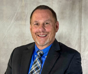 A smiling man with short hair and a trimmed beard, wearing a black suit jacket, blue shirt, and plaid tie, posed against a light gray background