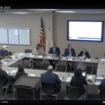 A group of people sit around a U shaped table in a conference room during the "ILA Meeting, June th " There is a large screen displaying a presentation, and an American flag is seen at the front
