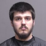 A young man with short dark hair, a mustache, and a beard is posing for a mugshot against a plain background, wearing a black garment typically used in mugshots
