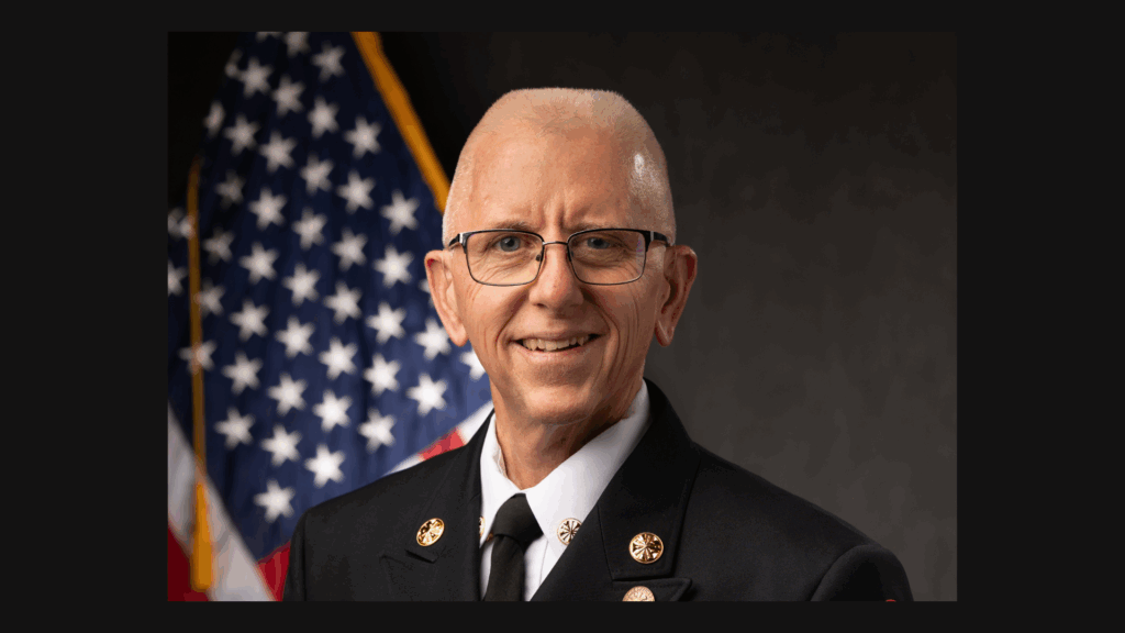 A smiling man wearing a formal dark uniform with gold insignia stands in front of the U S flag and a dark background He has short gray hair, glasses, and appears to be a fire department or public safety official