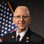 A smiling man wearing a formal dark uniform with gold insignia stands in front of the U S flag and a dark background He has short gray hair, glasses, and appears to be a fire department or public safety official
