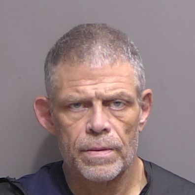 A middle aged man with short gray hair and a beard stares straight ahead with a neutral expression, wearing a dark colored shirt against a plain, light colored background