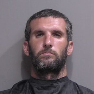 A man with short, dark curly hair and a beard is facing the camera against a plain gray background He is wearing a dark garment, possibly a drape or cape, around his shoulders
