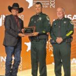 Three men stand on stage at the NSA Florida event One man in a cowboy hat and suit hands a plaque to a uniformed officer, while another uniformed officer stands beside them, all smiling for the camera