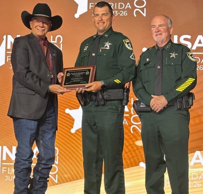 Three men stand on stage at the NSA Florida event One man in a cowboy hat and suit hands a plaque to a uniformed officer, while another uniformed officer stands beside them, all smiling for the camera