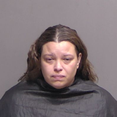 A woman with long brown hair and a neutral expression is wearing a black garment draped around her shoulders against a plain gray background