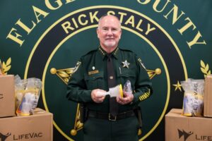 A uniformed law enforcement officer stands smiling, holding a LifeVac device He is surrounded by LifeVac boxes and packages, with a backdrop displaying "FLAGLER COUNTY" and "RICK STALY