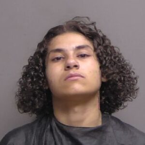 A person with curly, shoulder length dark hair is shown against a plain, neutral background, wearing a black garment Their expression is serious and they are looking directly at the camera