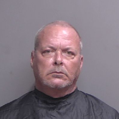 A middle aged man with short gray hair and a trimmed beard faces the camera with a neutral expression, wearing a black garment against a plain gray background