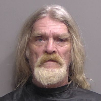 A man with long gray hair and a thick mustache and beard is photographed against a plain background, wearing a dark colored garment