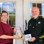 A woman and a uniformed sheriff stand smiling outside a building The woman holds a bag of evidence, while the sheriff holds a sign that reads "GIVING BACK EVIDENCE OF KINDNESS " A business hours sign is on the door behind them