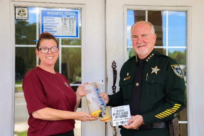A woman and a uniformed sheriff stand smiling outside a building The woman holds a bag of evidence, while the sheriff holds a sign that reads "GIVING BACK EVIDENCE OF KINDNESS " A business hours sign is on the door behind them