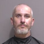 A middle aged man with short gray hair and a beard is shown in a mugshot against a plain gray background, wearing a black smock