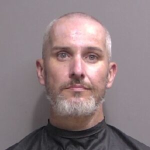 A middle aged man with short gray hair and a beard is shown in a mugshot against a plain gray background, wearing a black smock