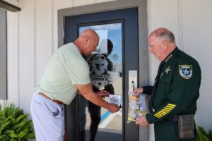 Two men, one in a light shirt and one in a police uniform, place a “Safe Place” sticker on a glass door The police officer holds a folder, and the door has a logo with a star and text