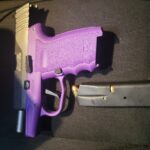 A semi automatic handgun with a purple grip and a black slide lies next to a detached black magazine loaded with a single visible bullet, all resting on a dark surface