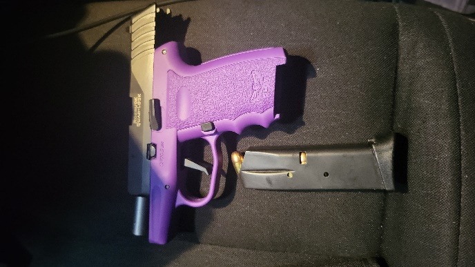A semi automatic handgun with a purple grip and a black slide lies next to a detached black magazine loaded with a single visible bullet, all resting on a dark surface