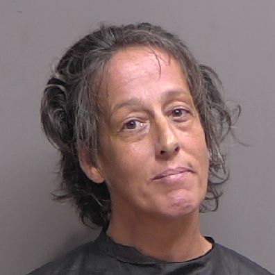A middle aged woman with short, wavy, graying hair looks at the camera with a neutral expression, set against a plain gray background She is wearing a dark, high collared garment