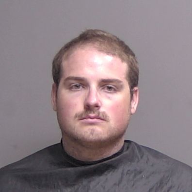 A young man with short light brown hair and a mustache is facing the camera against a plain gray background, wearing a dark drape over his shoulders