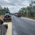Road construction site in Bunnell, FL, with heavy machinery, dump trucks, and piles of dirt along a freshly paved road Overcast sky and trees line both sides Text overlay displays date, time, location, and vehicle data