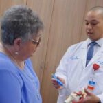 A doctor in a white coat explains a medical model to an older woman in a blue shirt during a consultation in a medical office