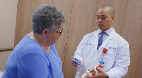 A doctor in a white coat explains a medical model to an older woman in a blue shirt during a consultation in a medical office