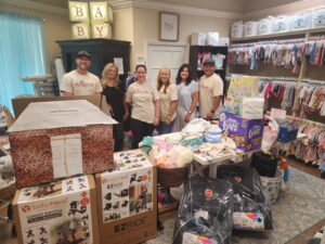 Seven people stand smiling behind tables filled with donated baby items—diapers, clothes, car seats, and more—in a room decorated with baby supplies and outfits A lighted "BABY" sign is visible above shelves of gifts