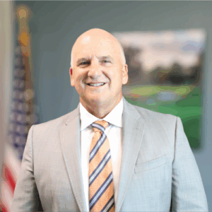 A smiling man in a light gray suit, striped tie, and white shirt stands indoors, with an American flag and a blurred painting in the background