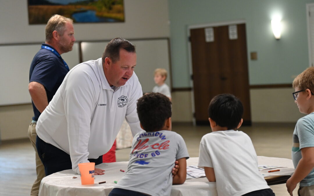A man in a white shirt leans over a round table, talking to three young boys who are standing around the table indoors Another adult stands in the background Art supplies are scattered on the table