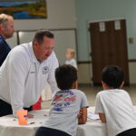 A man in a white shirt leans over a round table, talking to three young boys who are standing around the table indoors Another adult stands in the background Art supplies are scattered on the table