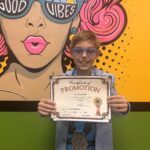 A smiling child with glasses holds a "Certificate of Promotion" in front of a colorful mural showing a woman with sunglasses that say "Good Vibes " The child wears a blue shirt and light jacket