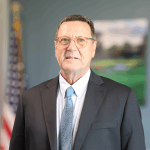 A man in a suit and tie stands indoors, with the American flag and a blurred framed picture in the background He has short brown hair, glasses, and a neutral expression A man in a suit and tie stands indoors, with the American flag and a blurred framed picture in the background He has short brown hair, glasses, and a neutral expression