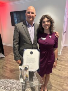 A man in a suit and a woman in a purple dress pose together indoors, smiling for the camera In front of them is an award and a framed certificate on a clear stand