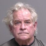 An older man with gray, curly hair and blue eyes poses for a mugshot against a plain gray background, wearing a dark collared shirt